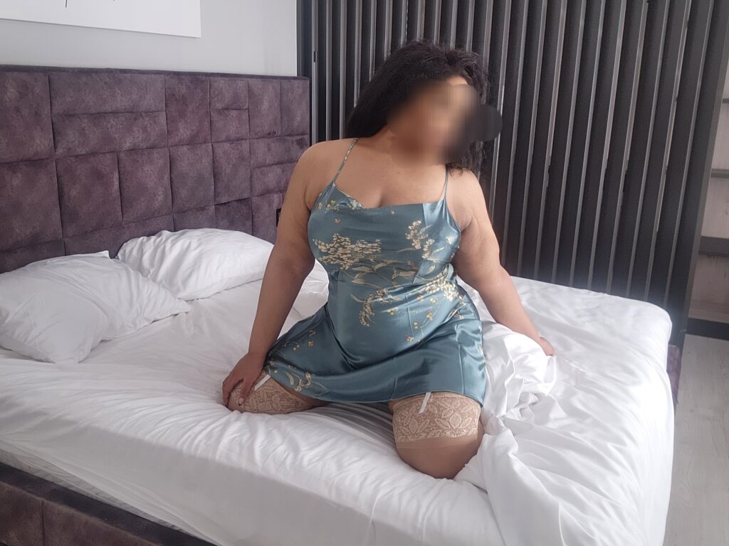 Busty overnight BBW escort London seated on bed in silk slip